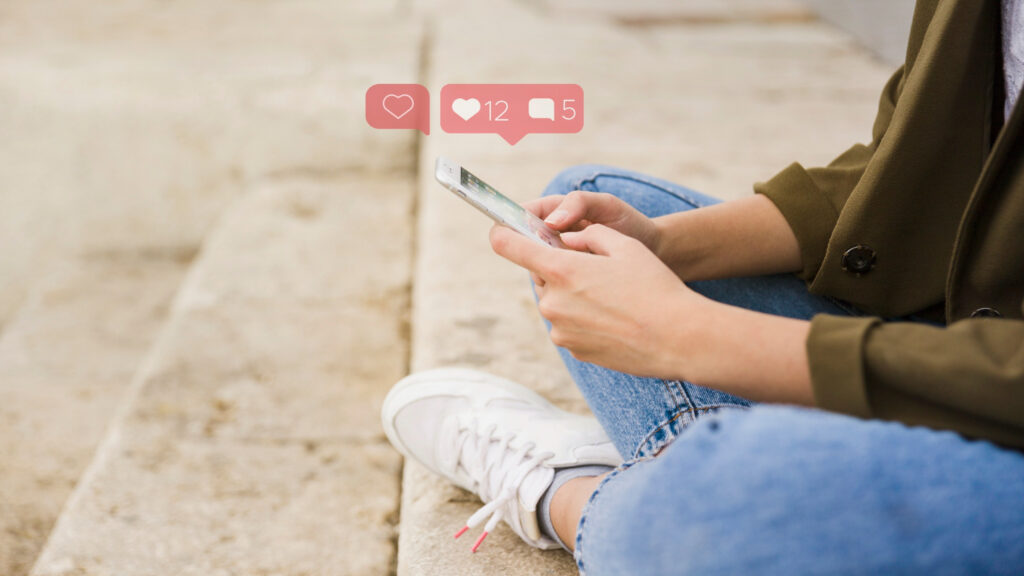 pessoas sentada em um degrau de escada utilizando um smartphone com ícones de mídias sociais suspensos na imagem