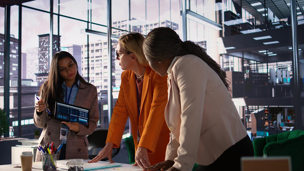 Custo de aquisição de cliente B2B: benchmarks e estratégias para a indústria 1 Custo de aquisição de cliente B2B: três mulheres executivas trabalhando em ambiente empresarial; uma delas segura um tablet.